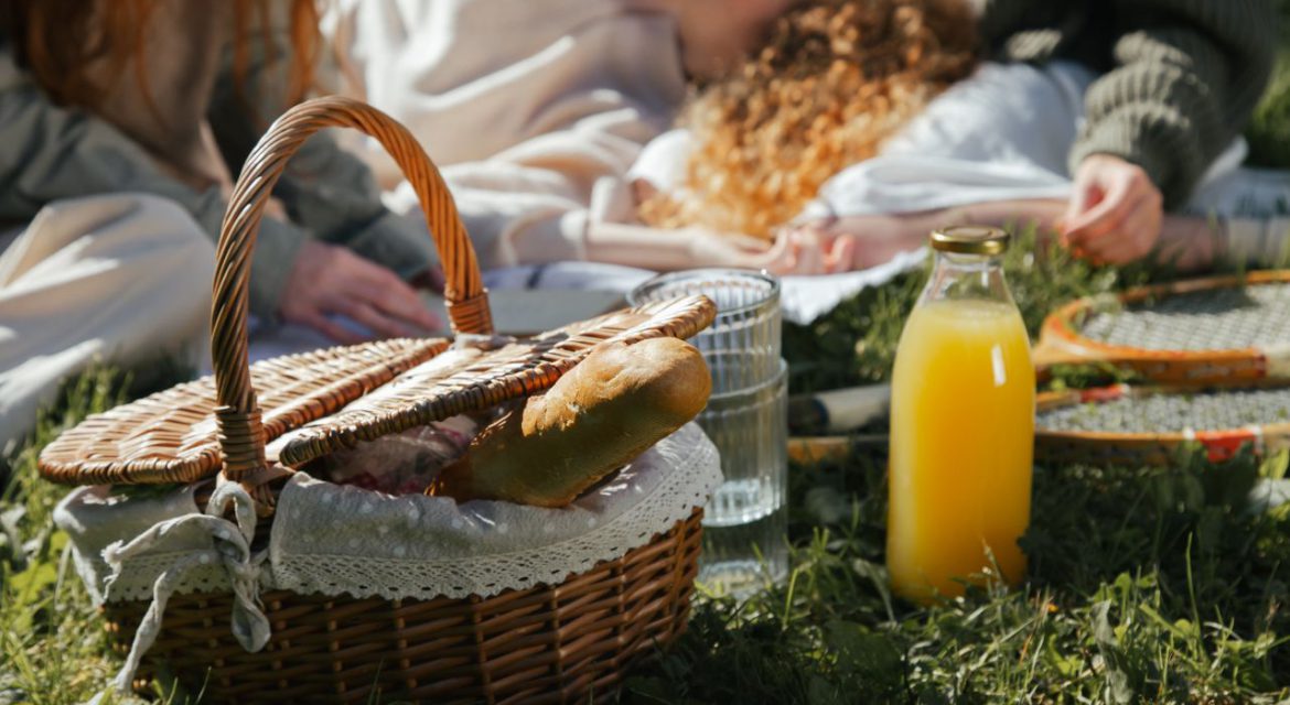 Picnic basket with bread, juice, and rackets on grass with people relaxing to illustrate a good picnic