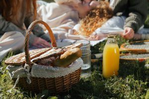 Picnic basket with bread, juice, and rackets on grass with people relaxing to illustrate a good picnic